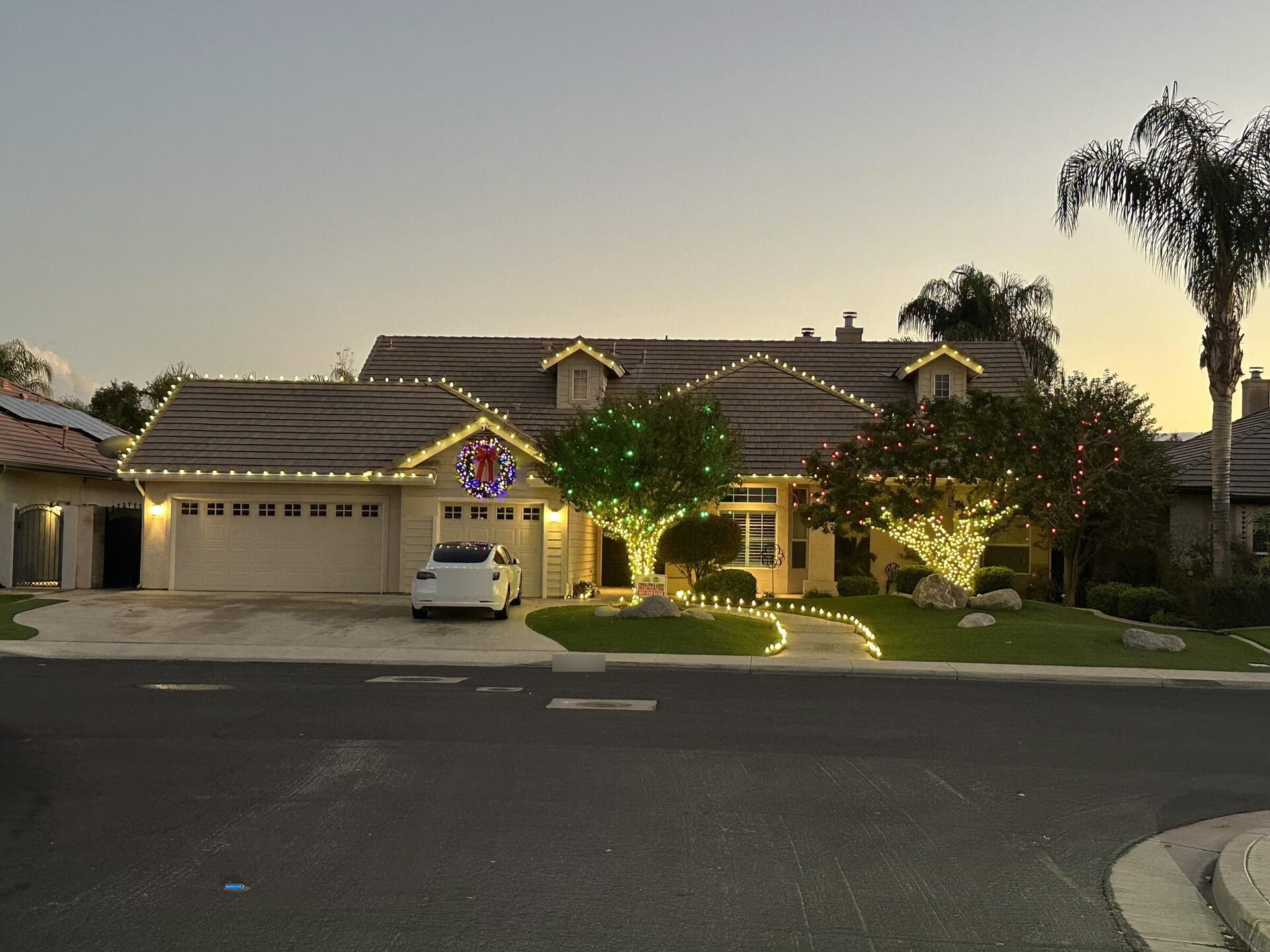 A Bakersfield home at twilight with warm white Christmas lights along the rooflines, a lit wreath and wrapped trees, installed by Just Be Bright